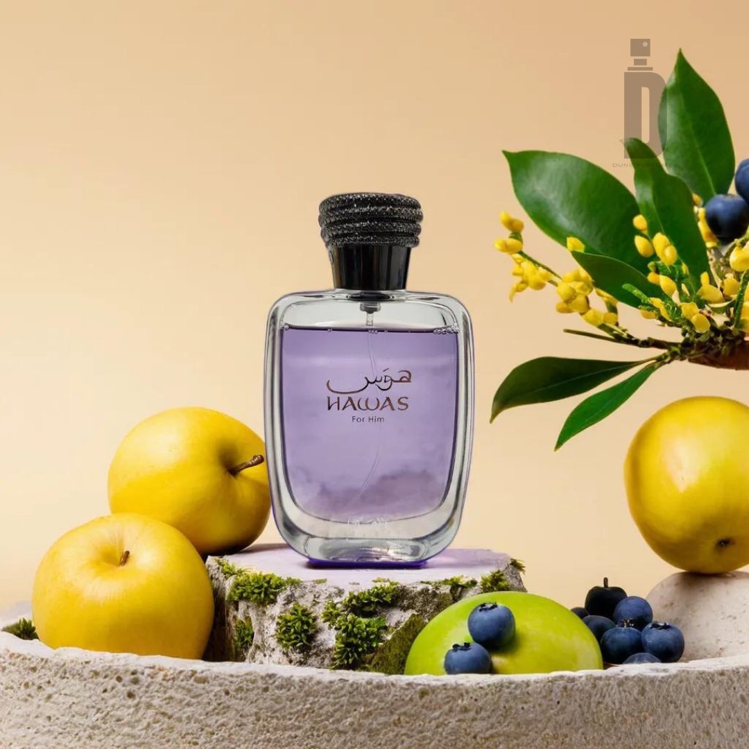 A glass bottle of "Hawas 100ml EDP - Rasasi" perfume with a black cap, filled with light purple liquid, is placed on a rustic surface. Surrounding the bottle are yellow apples, green apples, blueberries, and green leaves with small yellow flowers against a beige background, evoking adventure and allure.