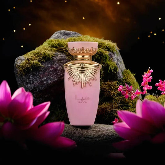 A bottle of Haya 100ml EDP from Lattafa, adorned in pink with gold accents, stands on a moss-covered rock. The backdrop showcases a starry night sky illuminated by a warm, glowing light. Pink flowers embellish the foreground, adding a touch of floral elegance to the scene and highlighting the sophisticated essence of Haya by Lattafa.