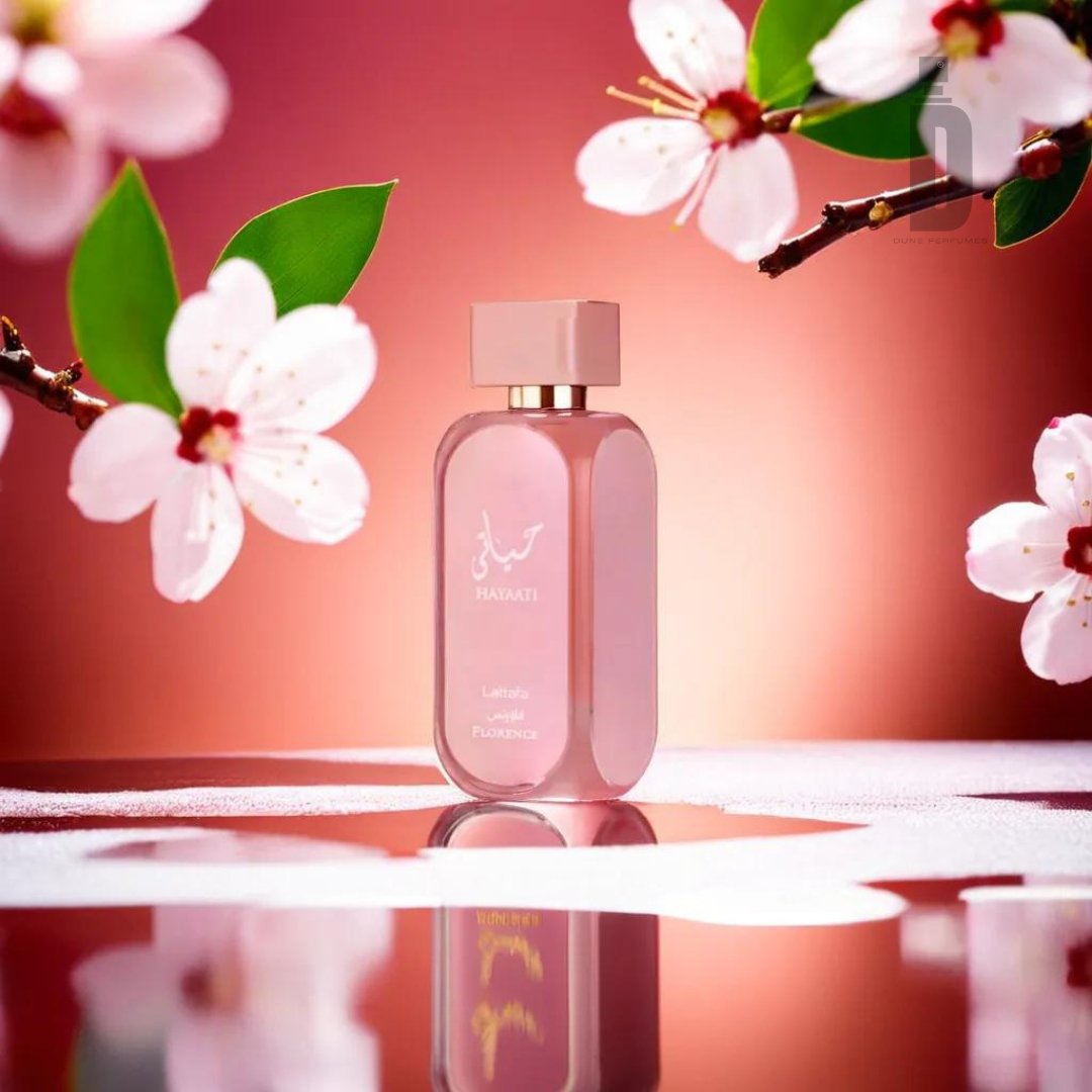 A pink perfume bottle labeled "Hayaati Florence 100ml EDP - Lattafa" is centered on a reflective surface adorned with water droplets. Surrounding the bottle are white cherry blossoms with green leaves, all set against a warm, red-orange gradient background. The scene conveys elegance, freshness, and exquisite Italian craftsmanship.