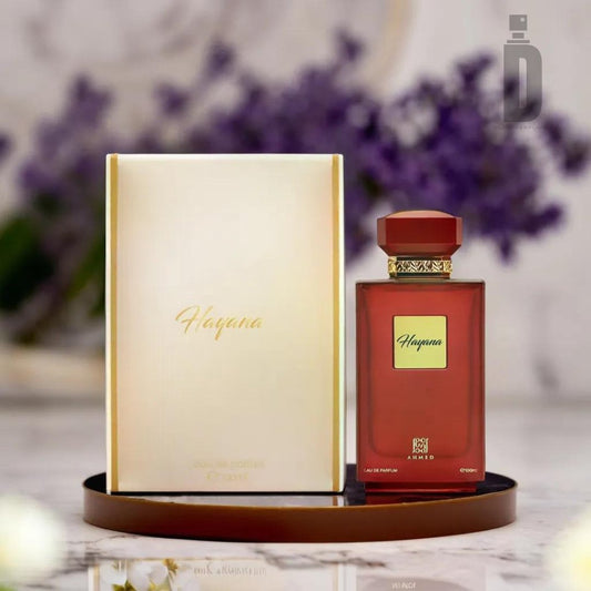 A bottle of Hayana 100ml EDP by Ahmed Al Maghribi is displayed alongside its matching box on a tray. The perfume bottle is red with a gold and red cap, and the box features a gradient white and gold design. A blurred floral backdrop adds an exotic charm to the scene, evoking a tropical escape.