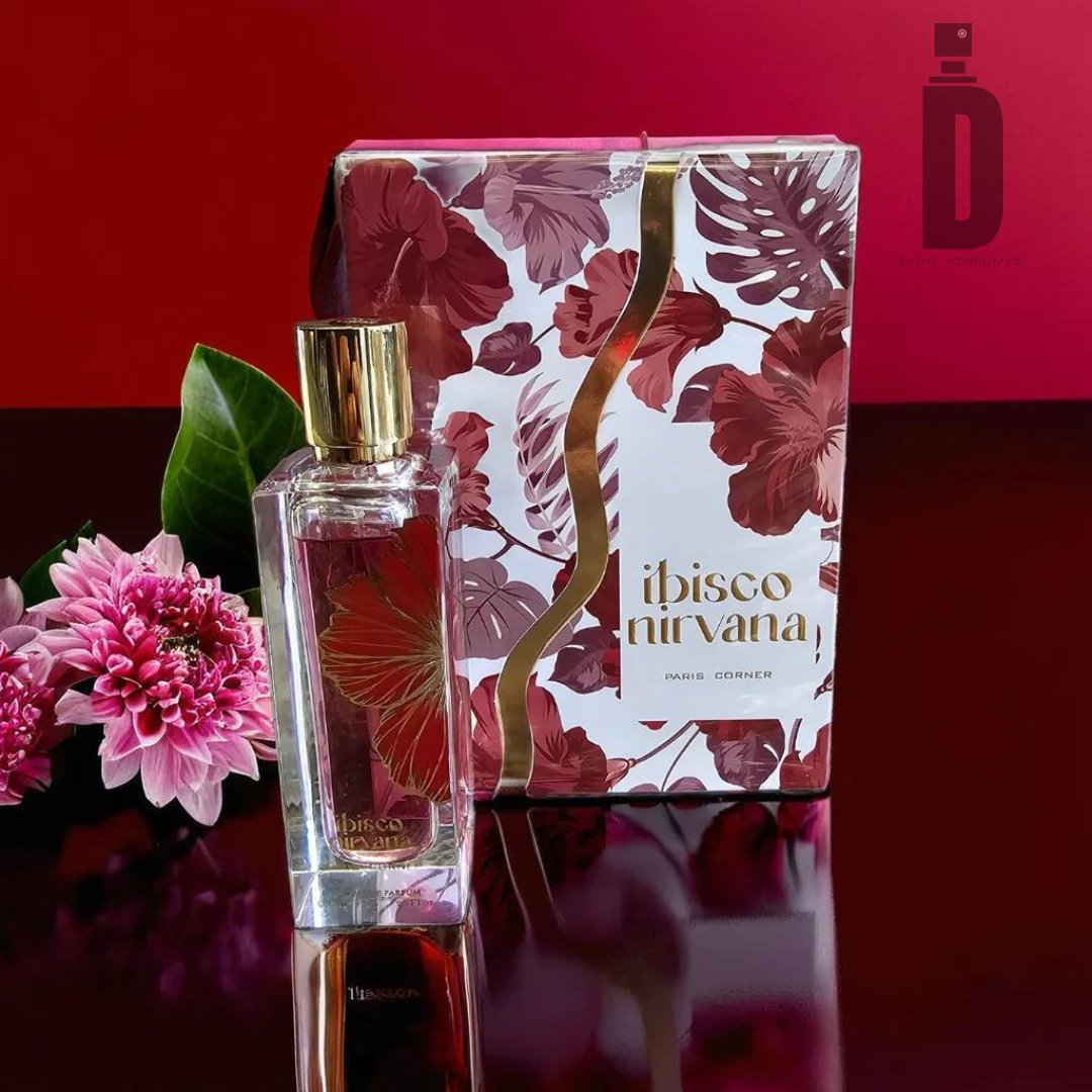 A 65ml bottle of Ibisco Nirvana EDP by Paris Corner is elegantly displayed in front of its floral-themed packaging. The sleek glass design, adorned with a gold cap, emanates luxury. Pink flowers are arranged next to the bottle, with a deep red background providing a rich contrast to the scene, reminiscent of Maison Crivelli Hibiscus Mahajad.