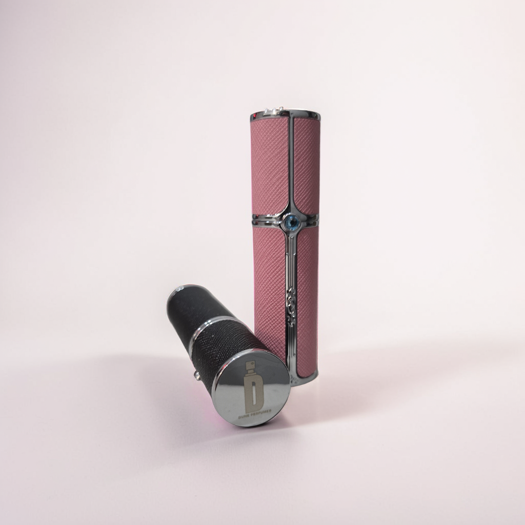 Two elegant, refillable Dune Perfumes travel sprays sit on a light pink background: one is a black and silver 10ml bottle; the other is a pink 5ml with metallic accents. Both are cylindrical with intricate detailing, perfect for travel convenience.