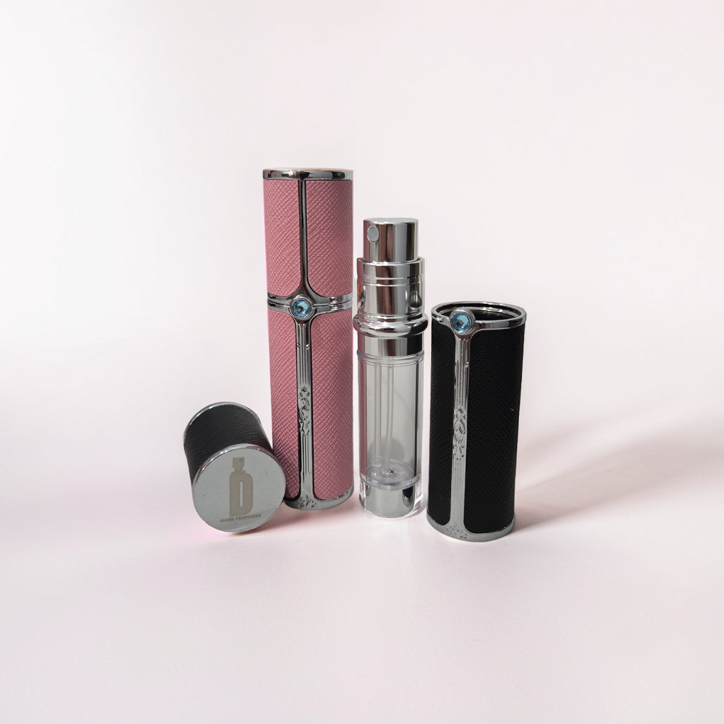 Two refillable perfume atomizers from Dune Perfumes stand upright on a light pink surface. The 5ml black atomizer is open, revealing a clear inner bottle, while the 10ml pink one is closed. Both have metallic accents and embossed designs, perfect for travel spray lovers.