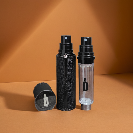 Two bottles from Dune Perfumes' 5ml & 10ml Travel Sprays are on a warm brown surface. One is a sleek black spray with texture, the other clear. A matching cap lies nearby, with light casting shadows.