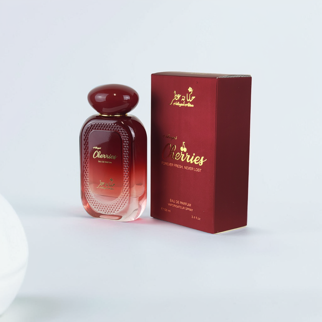 A red bottle of Cherries 100ml EDP by Hekayat Attar sits next to its matching box, both with gold text and embellishments. The fragrance has a subtle floral depth, elegantly highlighted by the simple white background.