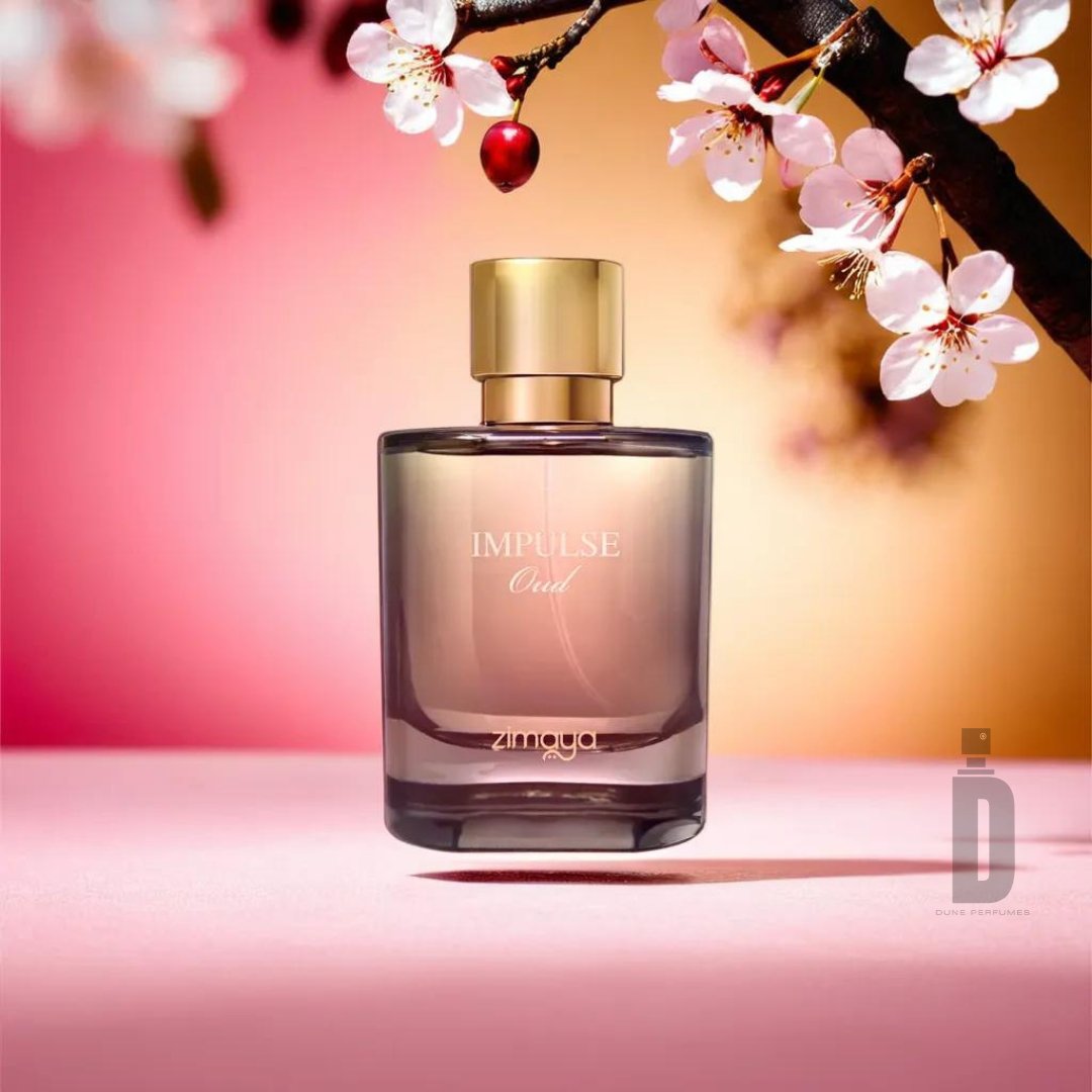 A perfume bottle labeled "Impulse Oud 100ml EDP - Zimaya" with a golden cap stands on a soft pink surface. Blooming cherry blossoms on a branch adorn the background, set against a gradient of pink, peach, and orange tones, creating an elegant ambiance that captures the sophisticated scent of this exquisite fragrance by Zimaya.