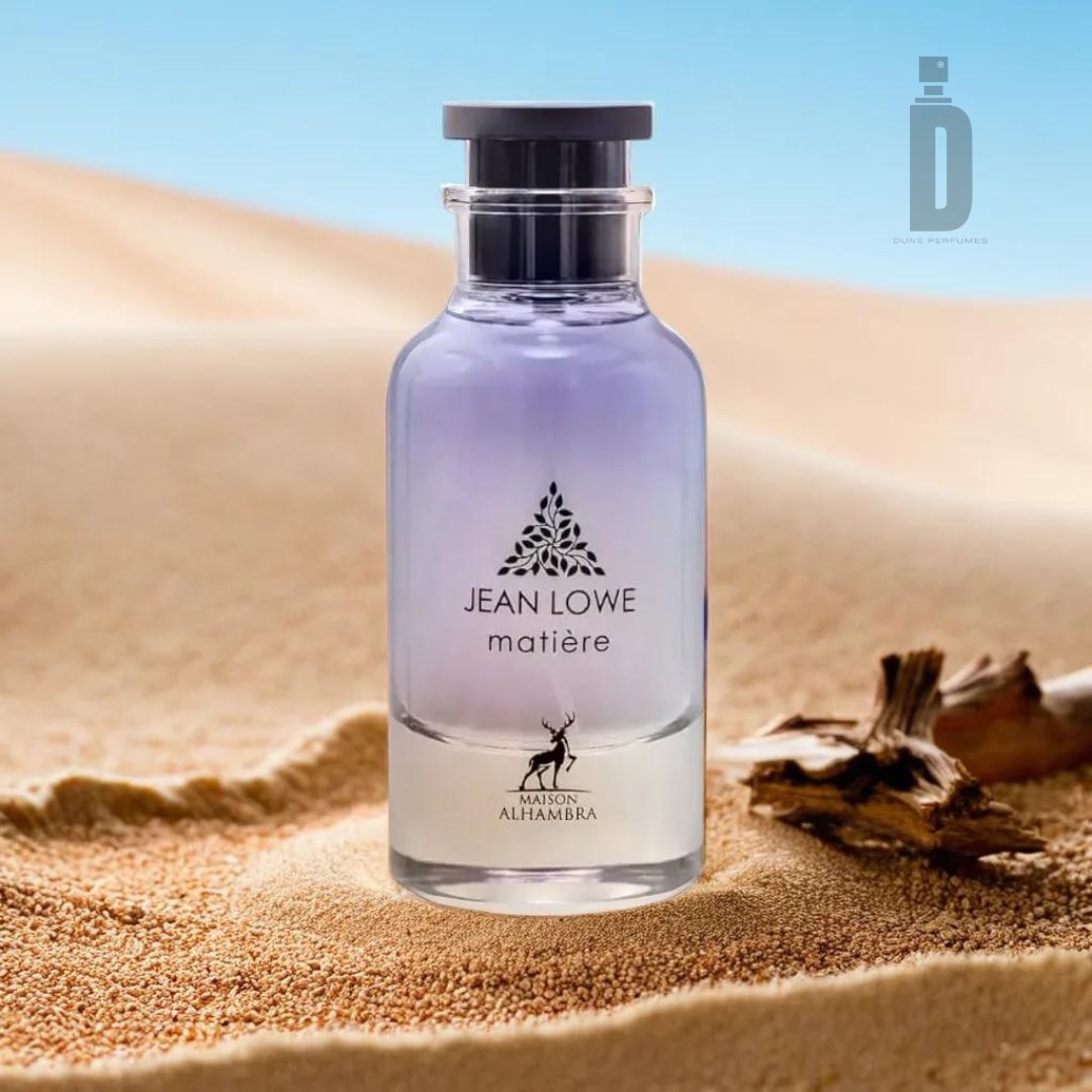 A 100ml EDP bottle labeled "Jean Lowe Maitre (Matiere)" by Maison Al Hambra is set against a desert background with sand dunes. The transparent bottle fades from light purple at the top to clear at the bottom, evoking olfactory artistry. A small piece of driftwood lies next to the captivating fragrance.