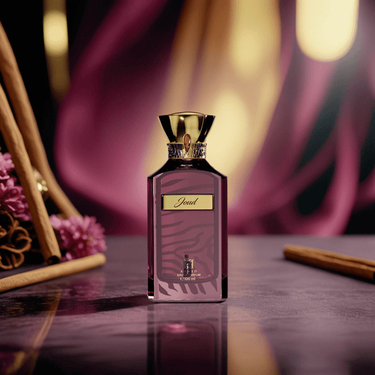 The Joud 100ml EDP by Ahmed Al Maghribi, featuring a gold-capped bottle and "Joud" label, rests on a reflective surface with flowers, cinnamon sticks, and vanilla, exuding a warm, spicy scent against a blurred purple and gold background.