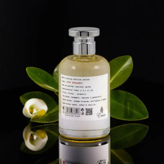 A bottle of parfum natural spray labeled "Just Bergamont 100ml EDP - Emir" by Paris Corner with a silver cap rests against a dark background. The fragrance stands on a reflective surface, surrounded by green leaves and small white flowers. The label lists scent notes and composition details, embodying Italian luxury.