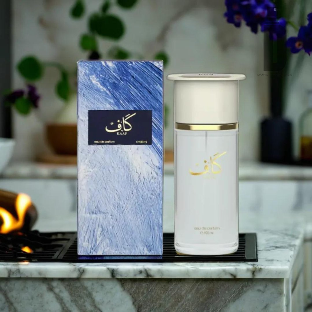 A beautifully designed Kaaf 100ml EDP - Ahmed Al Maghribi bottle with elegant Arabic script sits on a marble countertop next to its matching blue and white textured box. In the background, there is a blurred setting with a lit candle and purple flowers in a vase, adding to the elegant fragrance's allure.