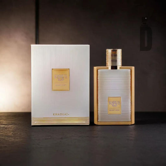 A bottle of Karus Secret Musk 100ml EDP by Khadlaj stands next to its matching rectangular box. The bottle and box have a white base with gold accents, and the name "Karus Secret Musk" is prominently displayed in the center. Soft lighting highlights the set, hinting at an exotic spice and Arabian tonka blend within.
