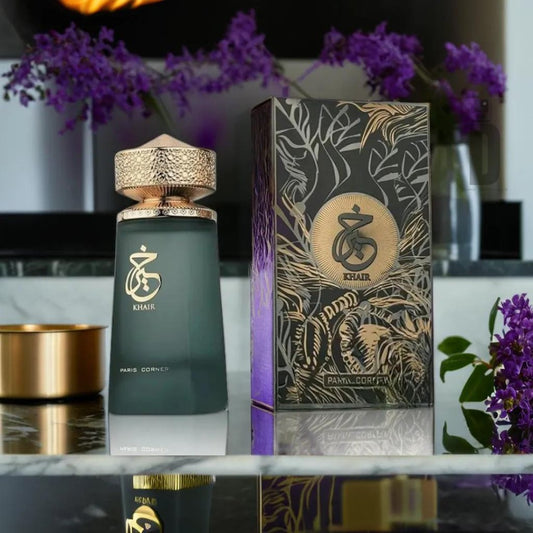 A perfume bottle labeled "Khair 100ml EDP - Paris Corner" stands next to an ornate box featuring the same branding. With a shiny cap and a gradient glass body, this Paris Corner fragrance exudes timeless elegance. Purple flowers and a lit candle in the background enhance the luxurious setting, evoking hints of Agarwood Oud.