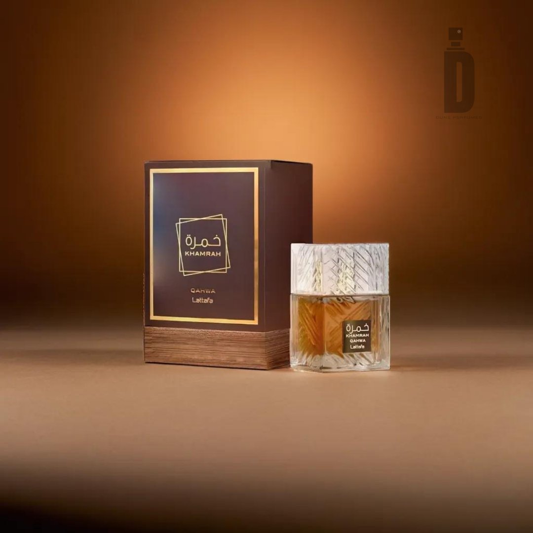 A bottle of Khamrah Qahwa 100ml EDP by Lattafa sits next to its brown and gold box against a gradient brown background. The clear bottle, with a textured glass design, contains amber-colored liquid. Radiating its captivating aroma, the packaging displays the brand "Lattafa" and product name "Khamrah Qahwa" in Arabic and English.