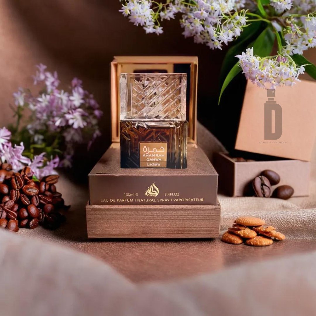 A bottle of Khamrah Qahwa 100ml EDP by Lattafa is displayed elegantly atop a stack of brown boxes. The background features delicate lavender flowers and a few coffee beans and almonds scattered around, enhancing the luxurious presentation with its captivating aroma.