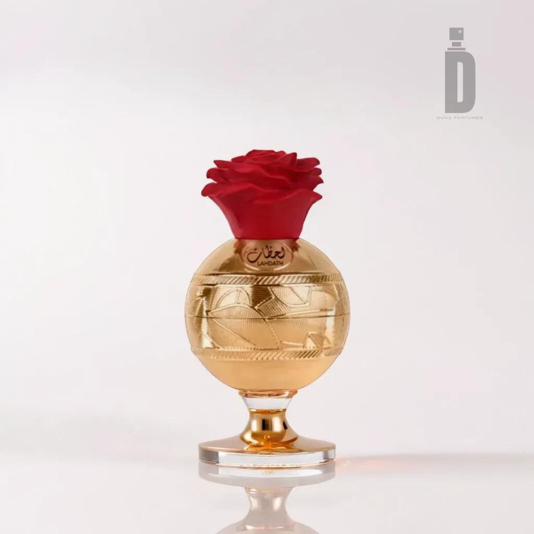 The Lahdath 80ml EDP - Lattafa is a luxurious perfume bottle with a gold-etched body and a red rose-shaped cap resting on a small pedestal base. Inspired by the timeless elegance of Lahdath fragrance, the bottle features an Arabic inscription near the top. A logo with a stylized letter "D" is visible in the top right corner.