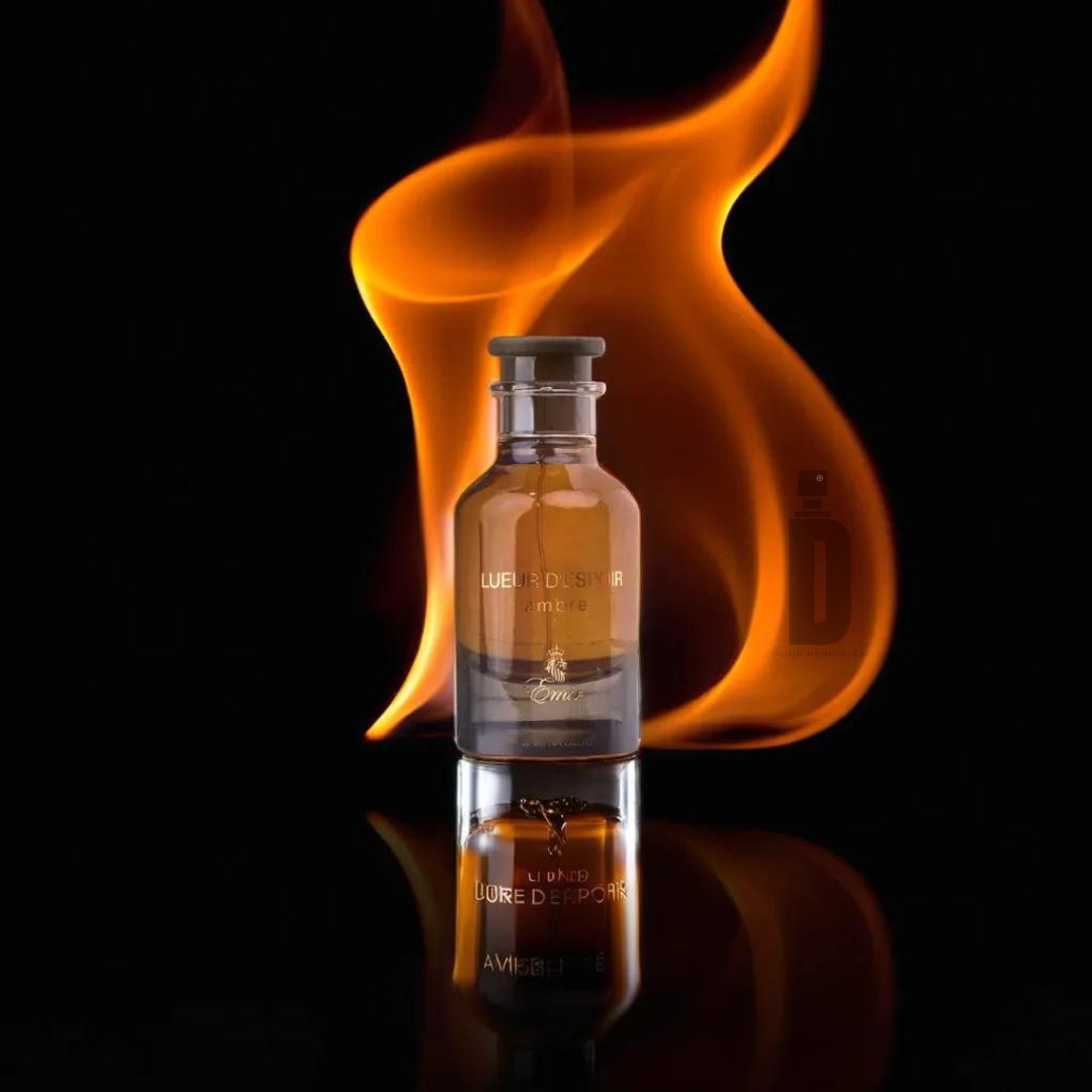 A glass perfume bottle labeled "Lueur D'espoir Ambre" by Paris Corner stands upright, its rich amberwood fragrance hinted at by an orange flame artistically rising behind it against a black background. The bottle's reflection is visible on the shiny surface beneath it.