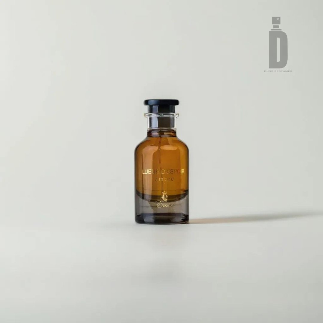 A small, dark brown glass perfume bottle with a black cap is centered against a light gray background. The label on the bottle reads "Lueur D'espoir Ambre" and indicates it's from Paris Corner, promising a refined amberwood fragrance that echoes the luxury of Lueur D'espoir Ambre 100ml EDP - Emir.