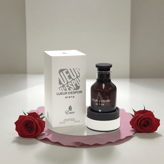 A 100ml bottle of Lueur D'espoir Arena EDP by Paris Corner stands next to its white box on a pink mat flanked by two red roses, evoking its luxurious rose and exotic oud notes, set against a neutral background.