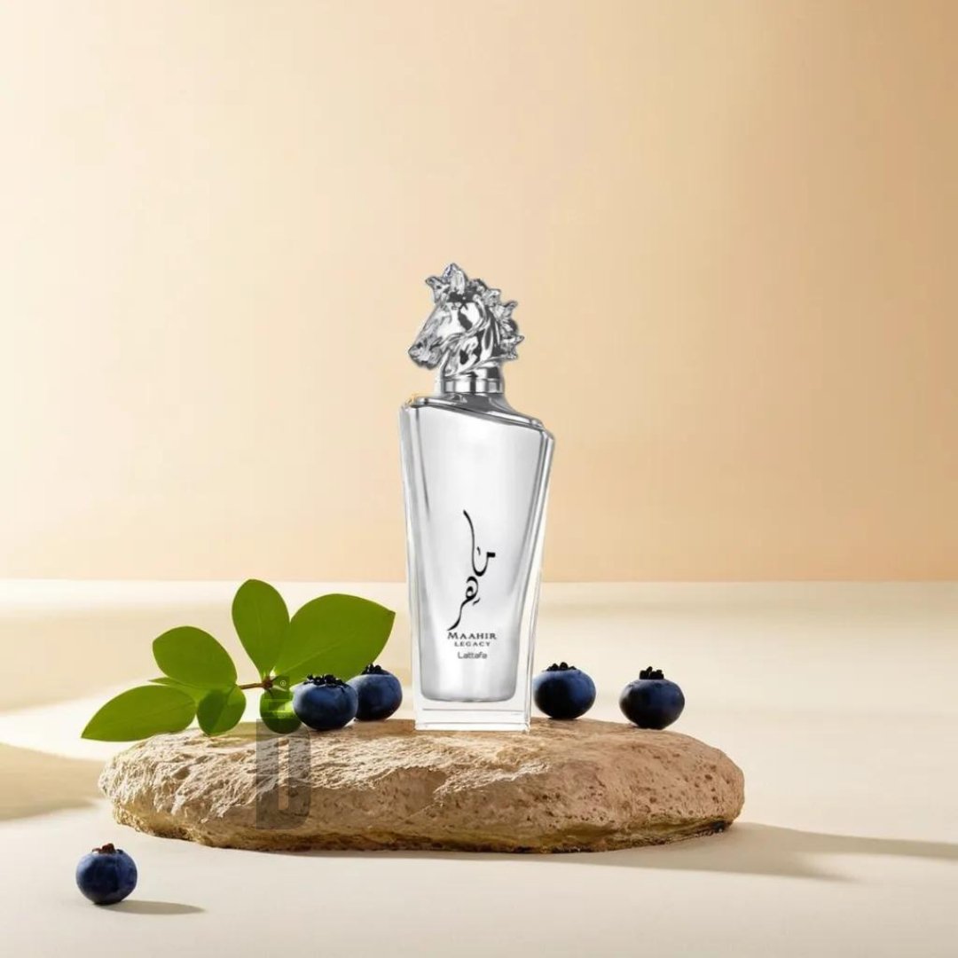 A sleek, transparent perfume bottle labeled Maahir Legacy 100ml EDP - Lattafa, with a sculpted, ornate metallic cap sits atop a textured rock. To the left of the rock is a small green plant, and scattered around are several blueberries, all set against a soft beige background. The sophisticated blend from Lattafa captures the essence of refined fragrance.