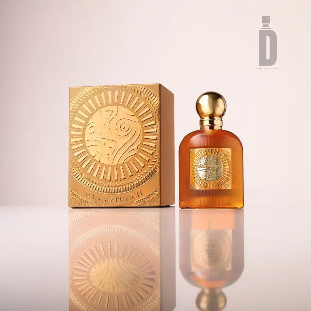 A bottle of amber-colored Mango Punch 100ml EDP by Emir from Paris Corner, featuring a gold spherical cap, stands next to a matching gold box, both adorned with a circular, embossed sun design. This tropical fragrance is set against a soft, neutral background that subtly reflects the elegant items.