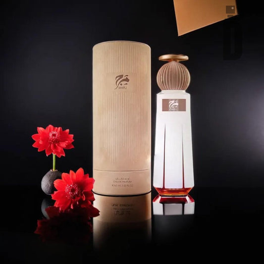 A frosted glass bottle of Marj 60ml EDP by Ahmed Al Maghribi, adorned with a decorative round cap, stands next to a cylindrical beige container. Red flowers are arranged beside the packaging, all set against a sleek black background with elegant lighting that highlights the complex fragrance of Marj EDP - Ahmed Al Maghribi.