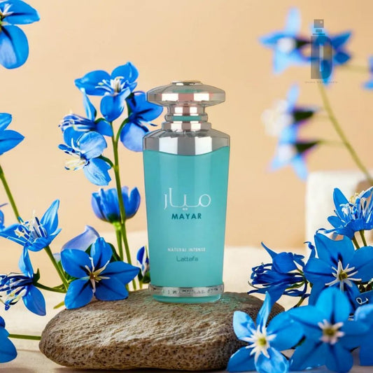 A blue perfume bottle labeled "Mayar Natural Intense 100ml EDP - Lattafa" by Lattafa stands on a flat rock, surrounded by vibrant blue flowers with green stems and leaves, capturing nature's essence fragrance against a subtle beige background. Another blurred perfume bottle is also partially visible in the background.