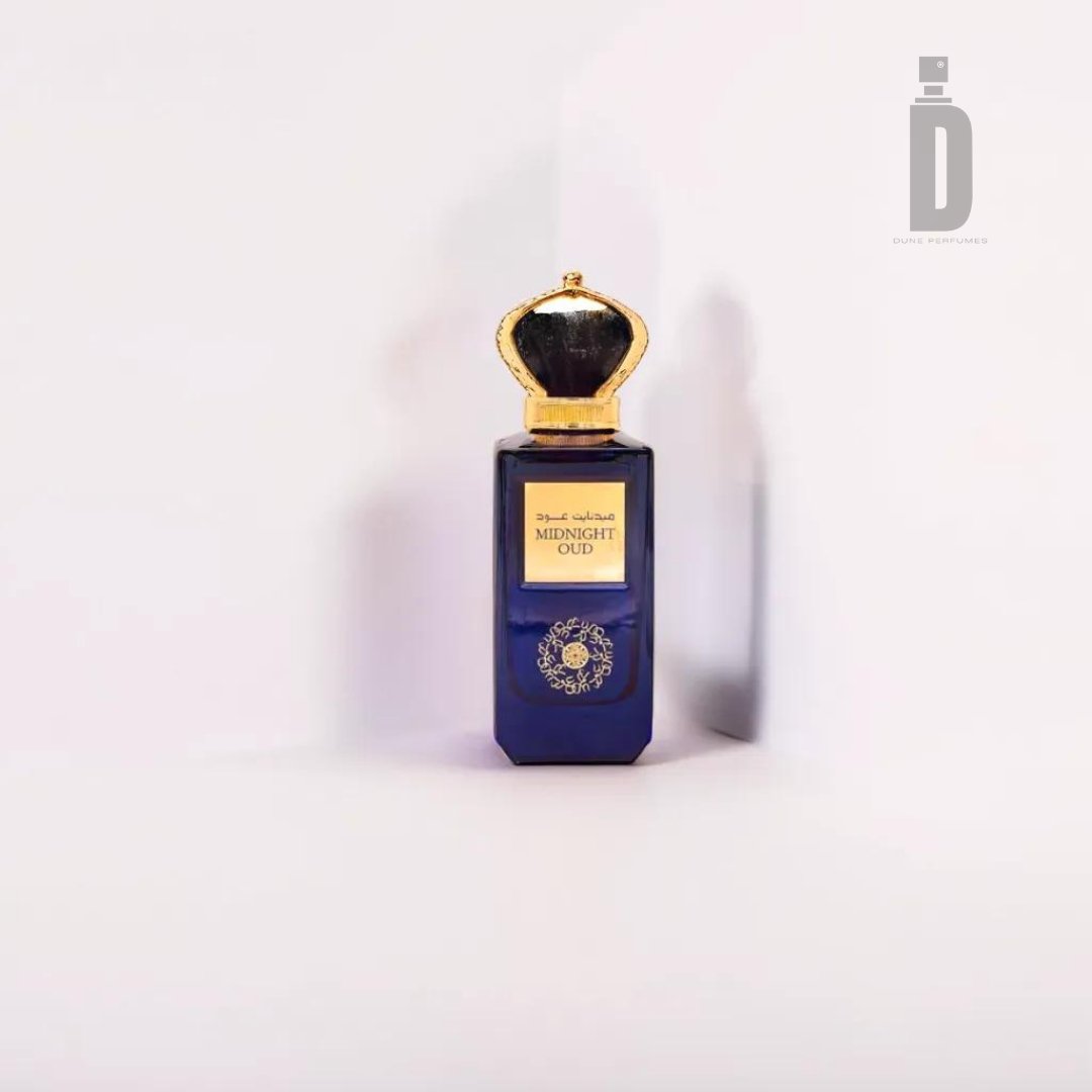A bottle of Midnight Oud 100ml EDP - Ard Al Zaafaran, a captivating fragrance from the brand Ard Al Zaafaran, is displayed against a plain white background. The bottle is navy blue with a gold cap and features a gold label with text in both English and Arabic. The logo for "Dar al Teeb," with a stylized "D" above it, is visible on the upper right.