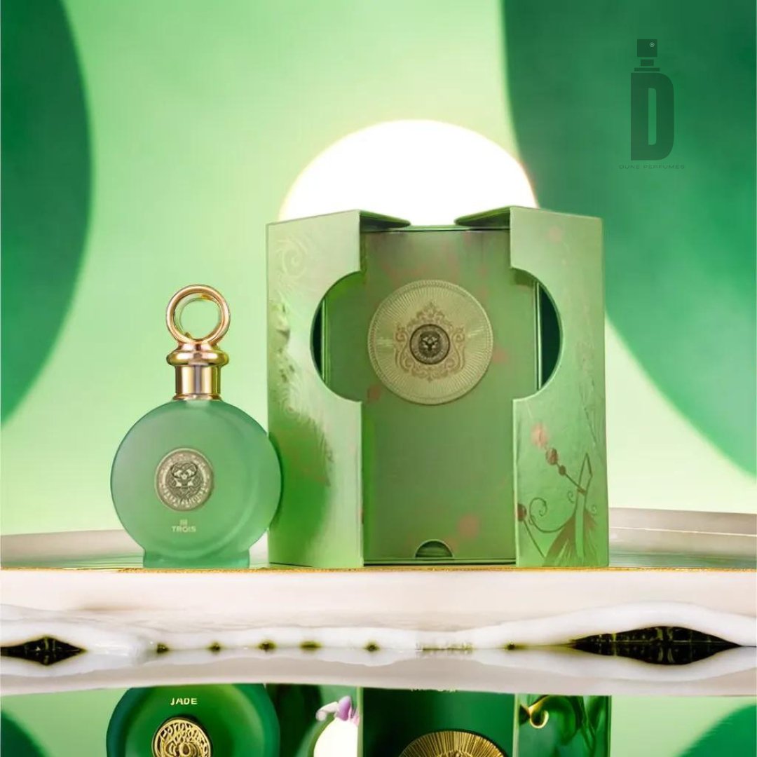 A green circular perfume bottle with a gold cap is placed next to an ornate green box adorned with intricate designs. The background, a gradient of green shades, creates a soft, elegant setting. The sophisticated fragrance, North Stag Expressions 3 Trois 100ml EDP by Paris Corner, reflects beautifully on the glossy surface below.