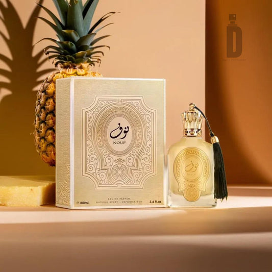 A Nouf 100ml EDP - Paris Corner bottle and its matching box are placed on a flat surface with a pineapple and a chunk of pineapple in the background. The elegant bottle, featuring a tassel attached to its neck, stands out alongside the intricately patterned box, perfectly capturing the essence of Nouf's signature fragrance style from Paris Corner.