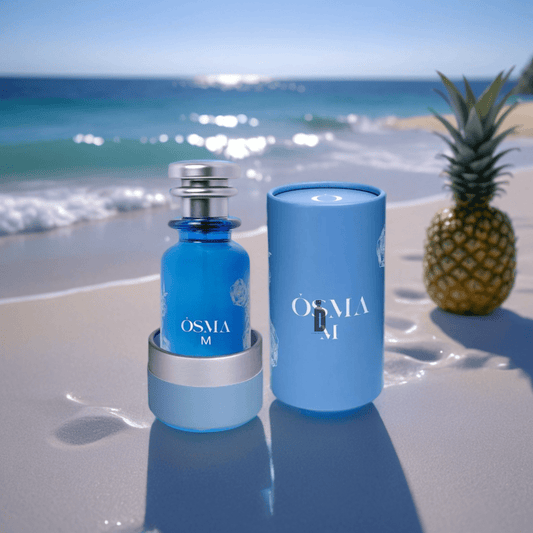 A blue Osma M 150ml EDP bottle with its box sits on sandy beach near the shore, beside a pineapple and coconut under a sunny sky.