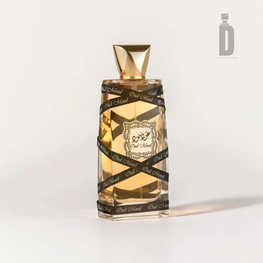 The Oud Mood 100ml EDP by Lattafa comes in a perfume bottle with a golden cap and a transparent glass body containing a light amber liquid. The bottle features black and gold decorative bands wrapped around it with text, and displays a logo with Arabic script on the front. Its exotic fragrance offers luxurious depth.