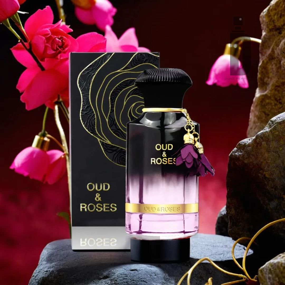 A luxurious bottle of Ahmed Al Maghribi's "Oud & Roses 60ml EDP" rests on a dark, rocky surface. The bottle is black at the top, seamlessly transitioning into a gradient of blush pink at the bottom. Pink flowers and the product box, also labeled "Oud & Roses," are in the background.