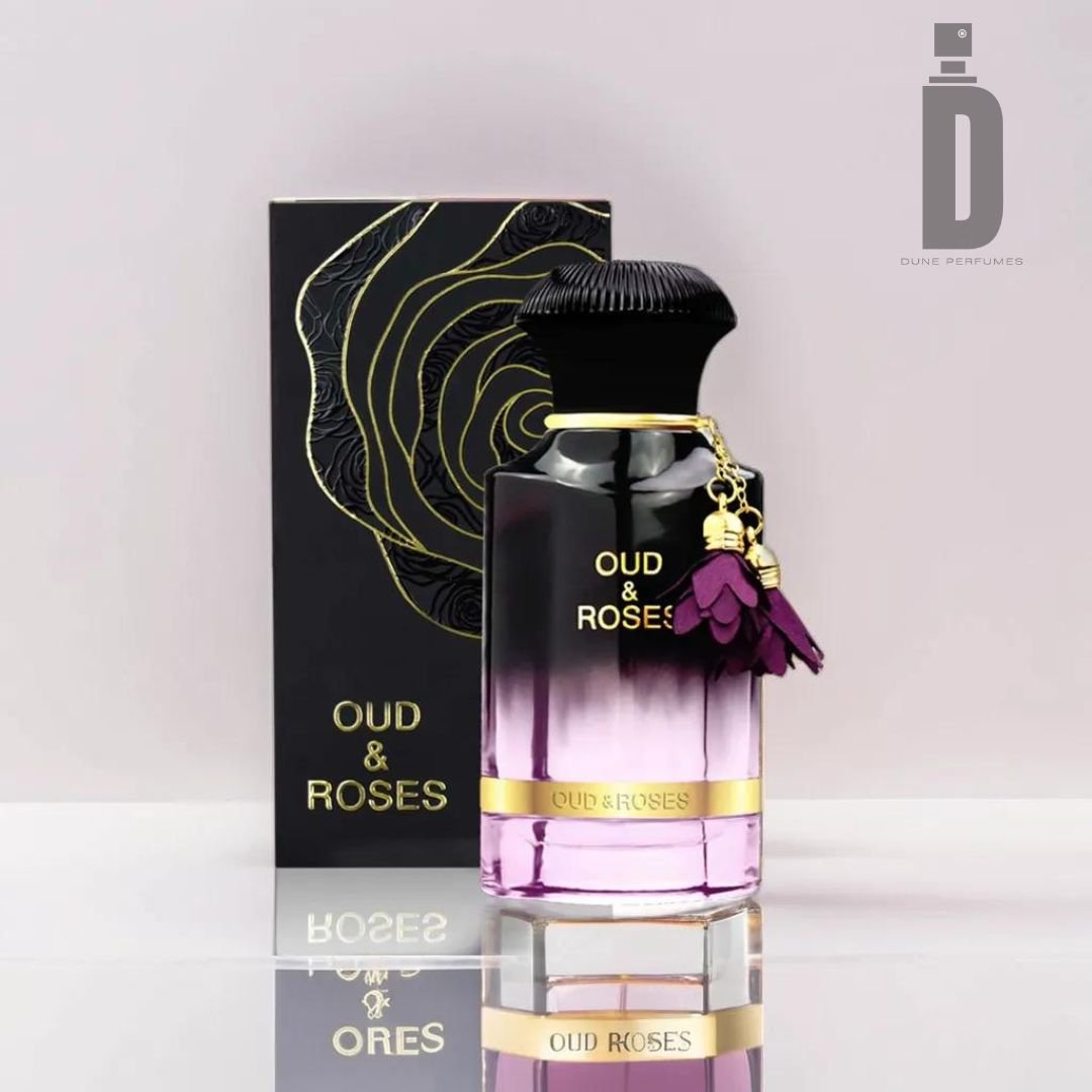 A black and pink bottle of "Oud & Roses 60ml EDP - Ahmed Al Maghribi" from the brand Ahmed Al Maghribi is placed in front of its matching box, which features a gold outline of a rose. The perfume bottle has a black cap adorned with purple flower charms, and the Dune Perfumes logo is visible in the top right corner.