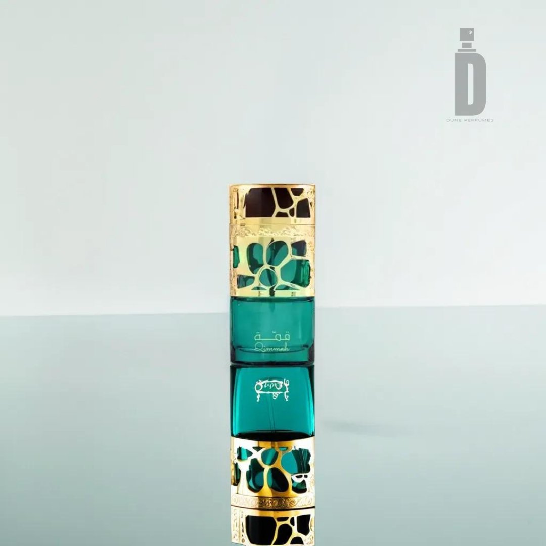 A green and gold perfume bottle with a decorative design sits upright on a reflective surface, showcasing its mirrored reflection below. Evoking femininity, it embodies the essence of Qimmah Women 100ml EDP by Lattafa. The minimalist, light-colored background highlights a logo with a stylized letter "D" in the upper right corner.
