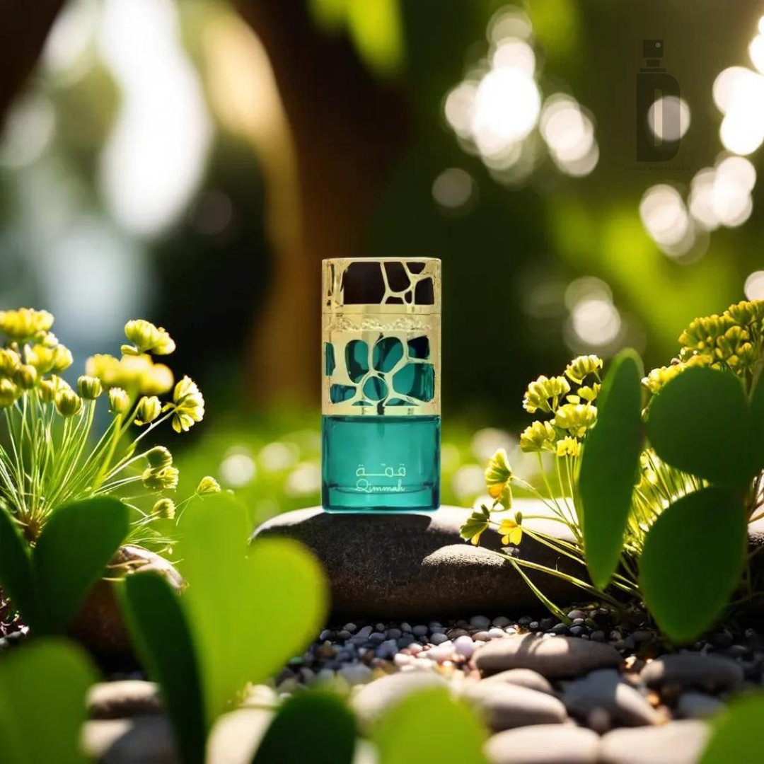A decorative Qimmah Women 100ml EDP - Lattafa bottle with a colorful, intricate cap is placed among small green plants and yellow flowers. Sunlight filters through the leaves, casting a warm glow on the scene. The fragrance bottle epitomizes femininity as it sits gracefully on a surface adorned with pebbles and leaves around it.