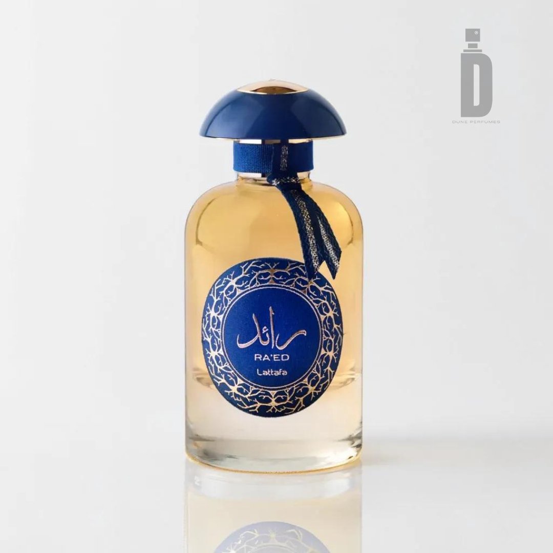 A glass perfume bottle filled with light golden liquid, featuring a blue rounded cap and a blue label adorned with intricate patterns and Arabic text. The label reads "RA'ED Lattafa." This luxury fragrance, Ra'ed Luxe Gold 100ml EDP by Lattafa, stands out against the white background, accented by a subtle logo in the top right corner.
