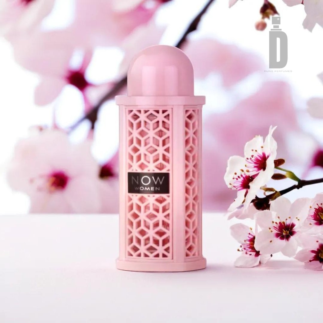 A pink, intricately designed perfume bottle with the label "Rave Now Women 100ml EDP - Lattafa" is positioned against a soft background featuring pink and white cherry blossom flowers. The minimalist and elegant presentation emphasizes the floral theme, warm undertones, and fruity notes of this feminine fragrance from Lattafa.