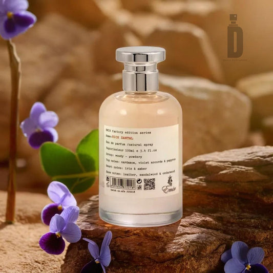 A bottle of Rich Santal 100ml EDP by Emir from Paris Corner, labeled with product details, sits on a rock. The background includes more rocks and small purple flowers, creating a natural, rustic setting that perfectly complements the sophisticated fragrance with notes of sandalwood.
