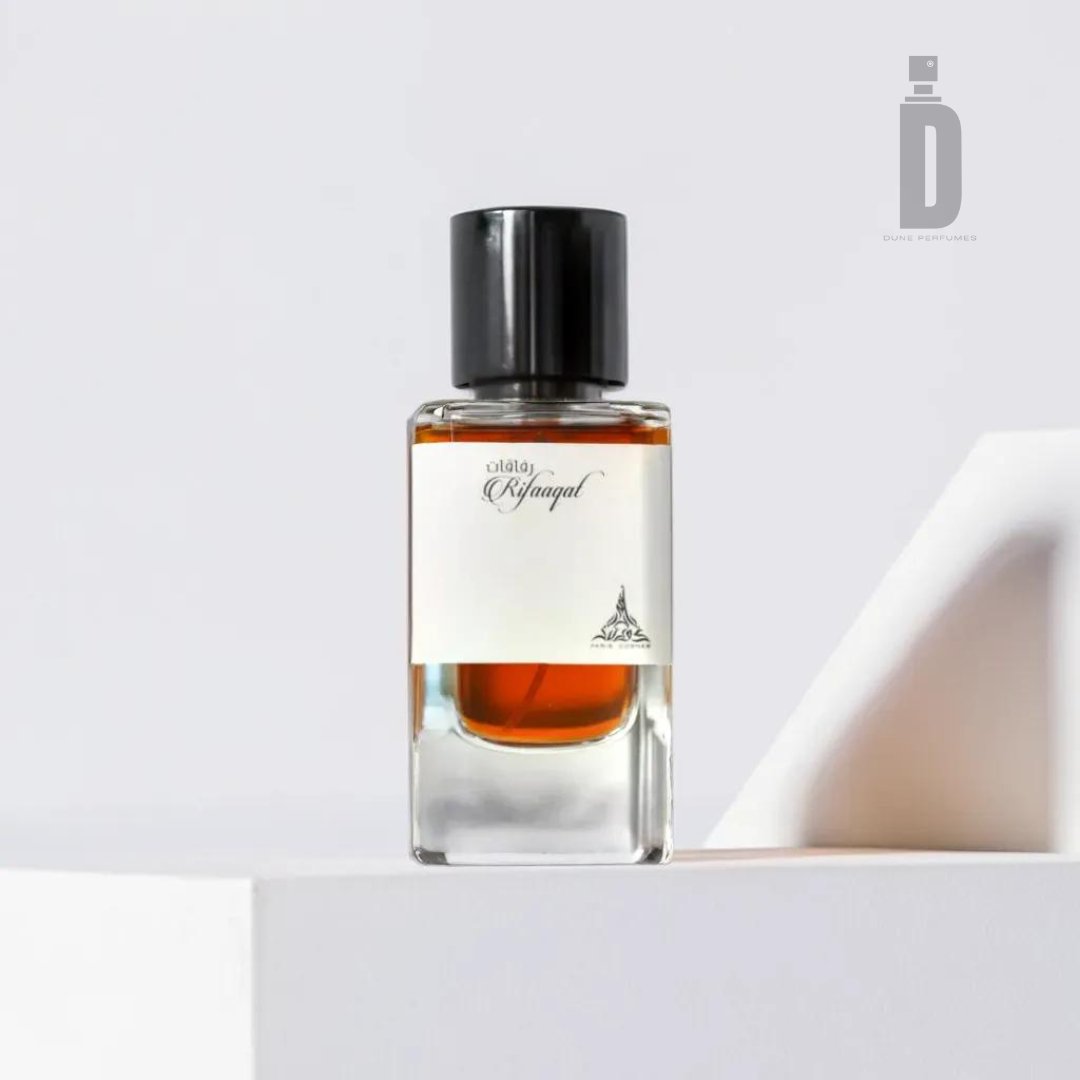 A minimalist bottle of cologne from Paris Corner with a black cap and a simple white label that reads "Rifaaqat 85ml EDP - Paris Corner." The amber-colored Rifaaqat fragrance is placed on a white platform with a light gray background, featuring a logo with a small, stylized bottle in the top-right corner.