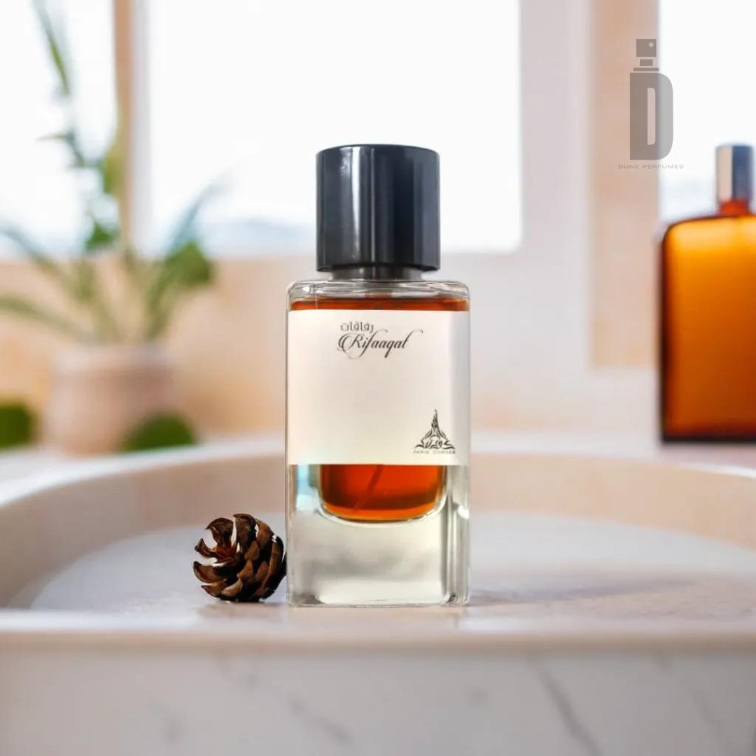 A bottle of cologne labeled "Rifaaqat 85ml EDP - Paris Corner" rests elegantly on a bathroom counter, embodying understated luxury. The bottle contains amber liquid and is topped with a sleek black cap. In the blurred background, another bottle and a green plant add to the serene setting. A small pine cone sits near the cologne bottle, enhancing its sophisticated aromatic ambiance.