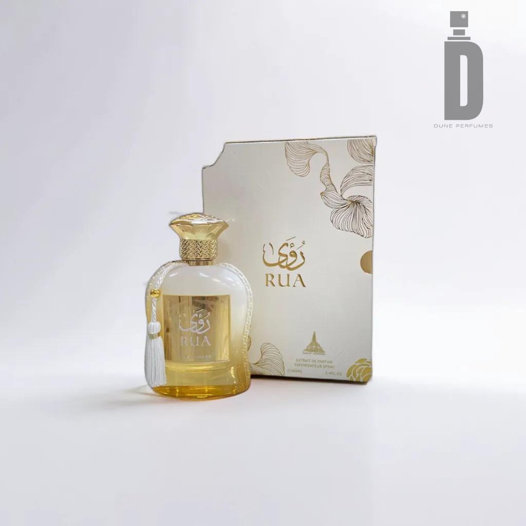 A bottle of Rua 100ml EDP - Paris Corner is placed next to its matching decorative box, exuding feminine elegance. The elegant bottle features a gold and white design with a delicate white tassel. The box showcases golden floral patterns and the Dune Perfumes logo, capturing the sophistication of Paris Corner.