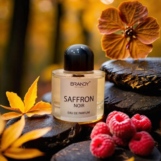 A bottle of "Saffron 100ml EDP" by Brandy Designs rests on dark stones with autumn leaves, vibrant pink flowers, and fresh raspberries surrounding it. The captivating aroma of this luxury perfume blends into the background's warm autumn colors, creating a cozy, seasonal ambiance.