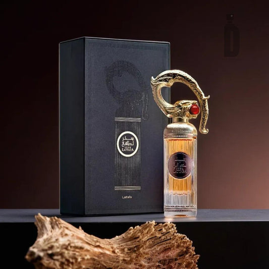 A perfume bottle with an ornamental, golden cap accented by a red jewel is displayed next to its matching black and gold box. The bottle contains an amber-colored liquid and stands on a dark surface. The box, labeled "Sehr Lattafa 100ml EDP - Lattafa," features an intricate design and logo, exuding luxury and sophistication.