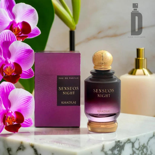 The "Sensous Night 100ML EDP - Khadlaj" bottle is showcased adjacent to its purple packaging on a marble surface. This alluring Eau de Parfum features a gradient purple design with a gold cap. In the backdrop, purple and white orchids and a gold dispenser enhance the presentation.