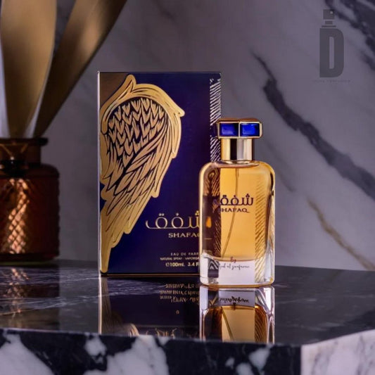 A bottle of Shafaq 100ml EDP - Ard Al Zaafaran is positioned in front of its box on a marble surface. The perfume bottle, evoking an exotic allure, has gold detailing and a blue cap, while the box features a gold wing design. An ornate vase is visible in the background.