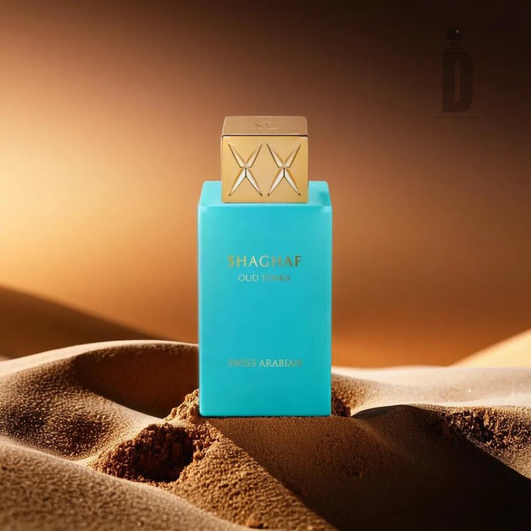 A turquoise bottle of Shaghaf Oud Tonka 75ml EDP by Swiss Arabian sits on sandy dunes, with a gradient brown background creating a warm, desert-like atmosphere. The bottle, adorned with a geometric patterned golden cap, hints at its exotic blend of Turkish Rose and Cinnamon Bark notes.