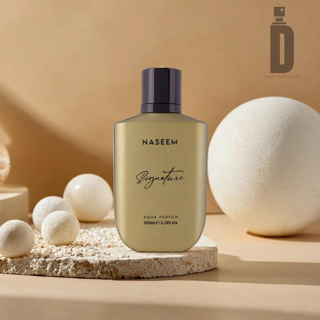 A rectangular bottle of Signature Golden Aqua Parfum by Naseem Perfumes (Alcohol-Free), 100ml/3.3fl oz, sits on a textured white surface. The background features minimalistic, round cream-colored decor pieces and a soft beige backdrop, with the Dune Perfumes logo in the upper right corner. This alcohol-free fragrance promises to be long-lasting.