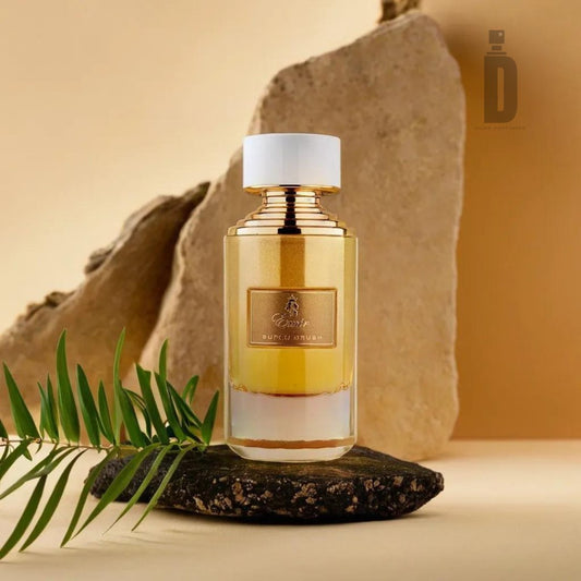 A stylish bottle of Super Crush 100ml EDP from Paris Corner rests on a dark stone, adorned with a white cap and framed by lush green leaves. Behind the bottle, large beige stone slabs provide an elegant and natural backdrop. The gold label with intricate details suggests the irresistible allure of its captivating fragrance.