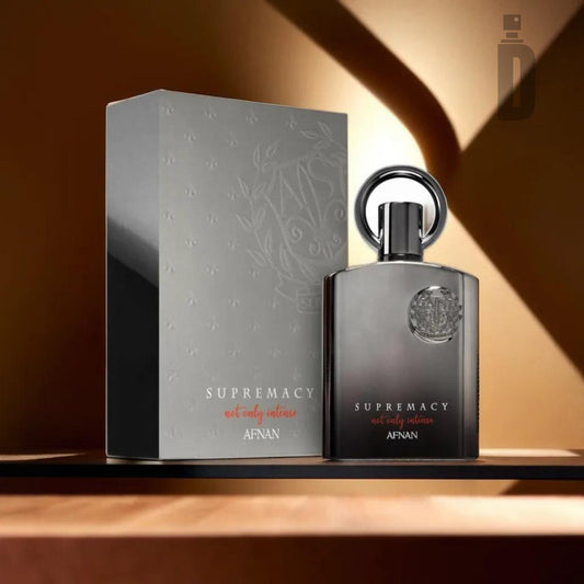 Introducing an image of the Supremacy Not Only Intense 100ml EDP by Afnan. This exquisite fragrance is housed in a silver-accented black bottle with a distinctive round cap, exuding sophistication. It is perfectly complemented by its dark gray box. The warm, dramatic lighting in the background accentuates the refined blend inside.