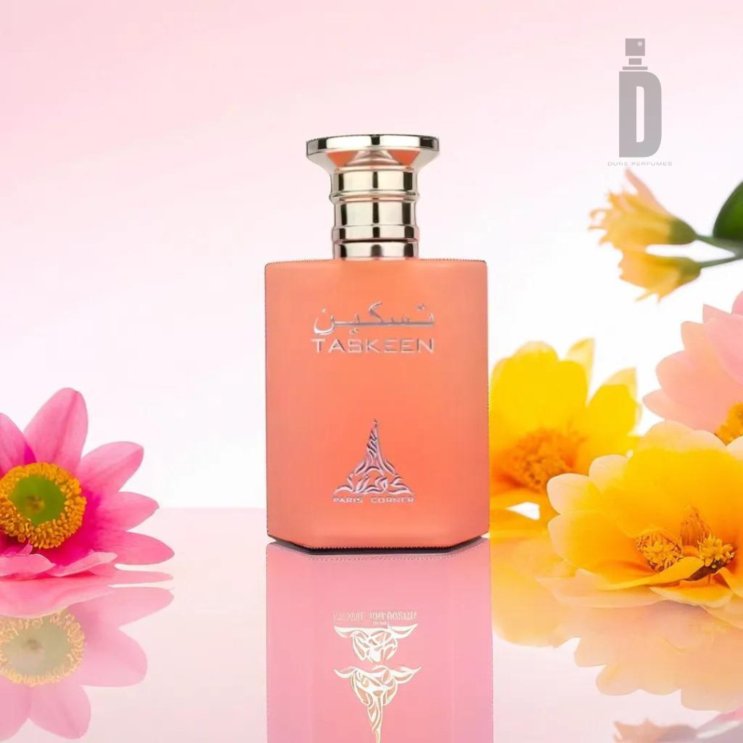 A pink and orange Taskeen 100ml EDP - Paris Corner perfume bottle with a silver cap and Arabic text stands amidst vibrant pink, yellow, and orange flowers on a soft pink gradient background. The Paris Corner logo featuring "D" is in the top right corner. The bottle's reflection is visible on the glossy surface, showcasing its luxurious fragrance.